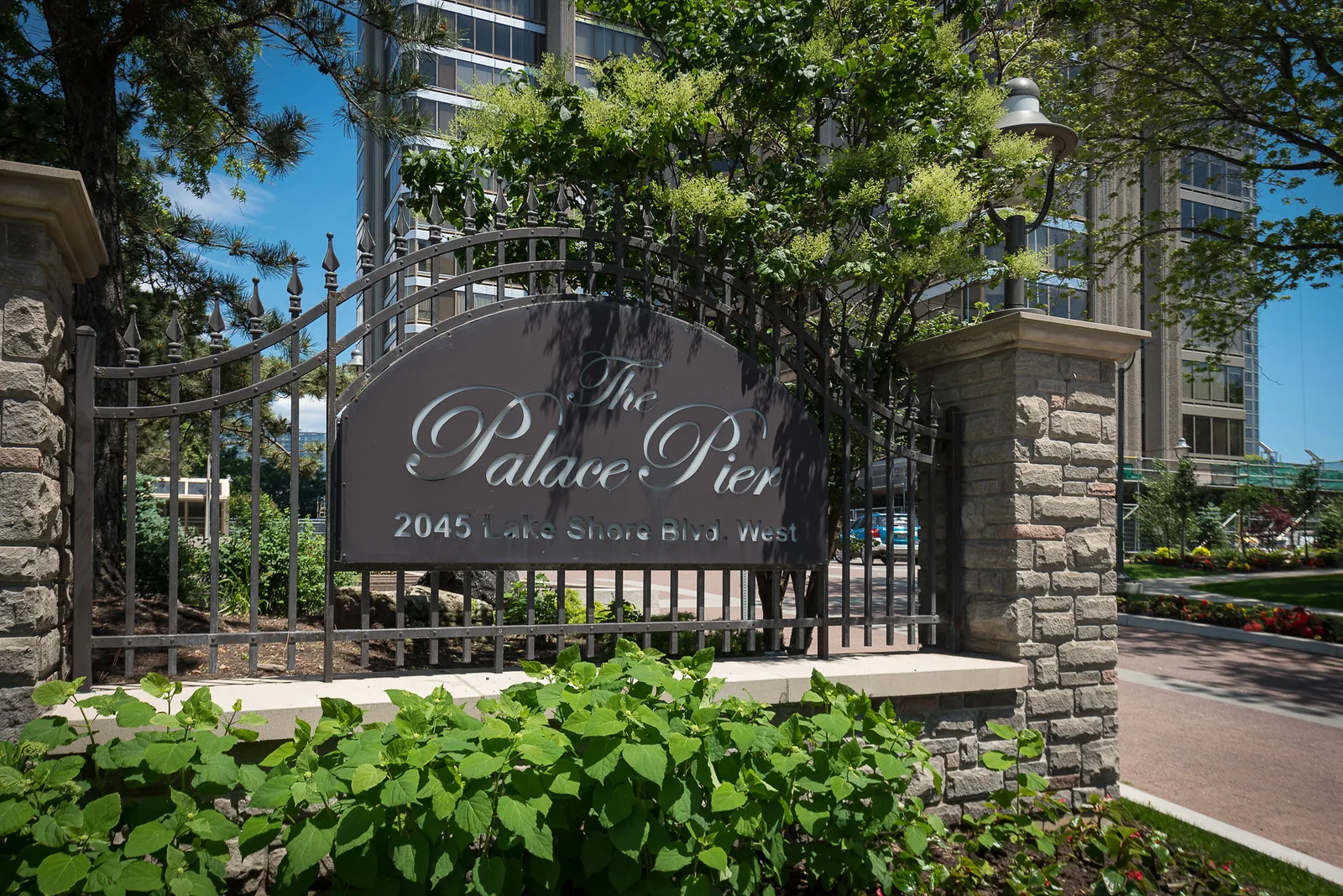 The stone and wrought-iron entrance sign for The Palace Pier at 2045 Lake Shore Blvd. West, framed by trees with the residence tower behind